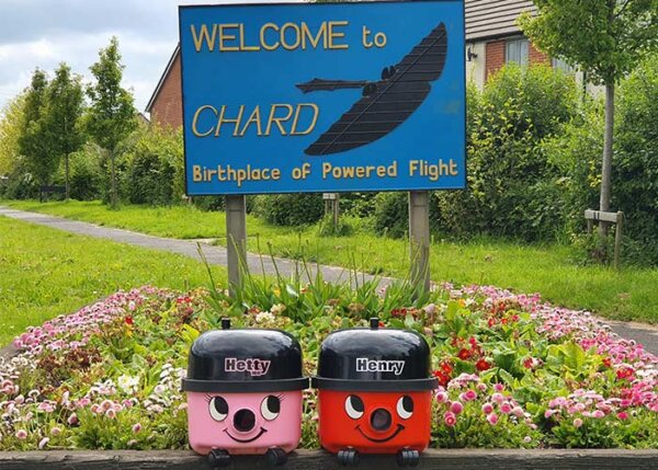 Numatic Red Henry and Pink Hetty in front of Welcome to Chard sign with a lush green backdrop